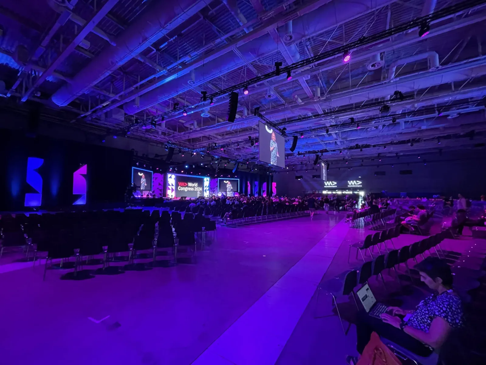 A wide view of WeAreDevelopers World Congress 2024, with a large stage, purple and blue lighting, and attendees watching a talk.