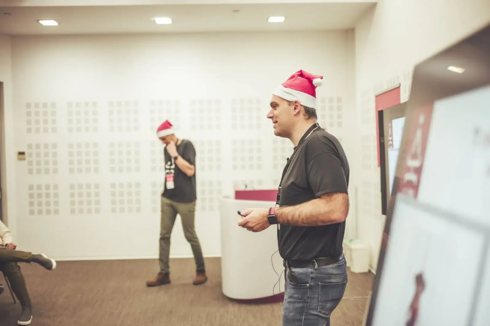A speaker wearing a Santa hat presents during a developer event, standing beside a large screen while another speaker waits in the background.